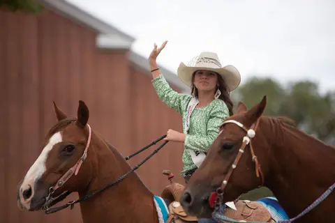 A young cowgirl wearing a cowboy hat and sash waves while riding a horse.