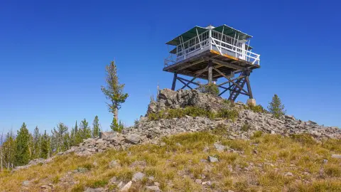A fire lookout tower stands atop a rocky mountain peak against a clear blue sky.