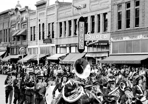 A marching band and crowd line a street with storefronts.