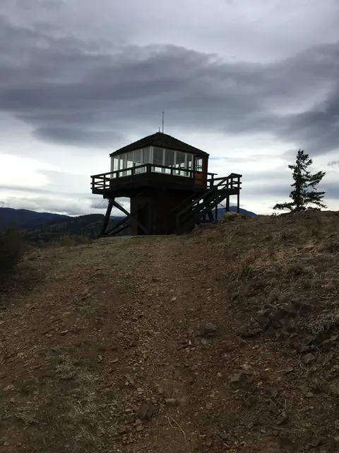 A fire lookout tower stands on a hill under a cloudy sky.