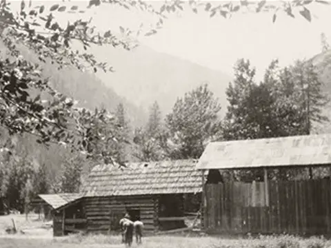 A historic photo shows a rustic log cabin and shed in a wooded mountain setting.