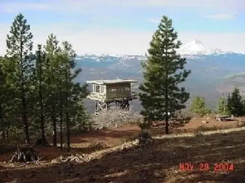 A fire lookout tower stands among pine trees in a mountainous landscape.