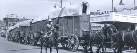 Black and white historic photo showing covered wagons and horses on a street.