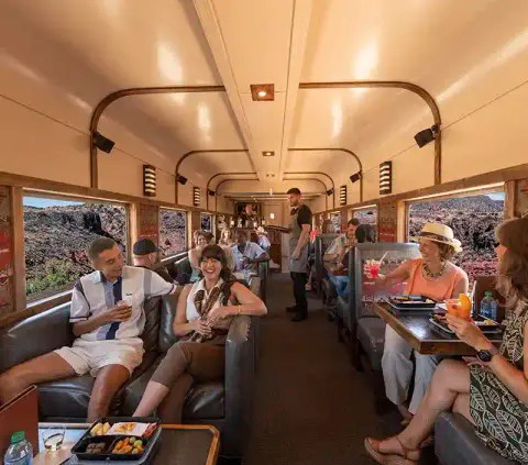 Passengers dine and relax inside a train car with large windows overlooking a rocky landscape.