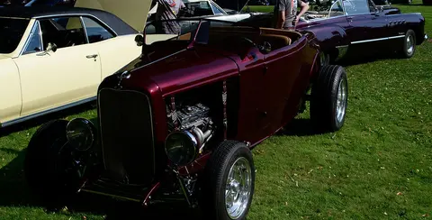 A dark red vintage hot rod is parked on the grass at a car show.