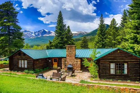 A log cabin with a stone chimney and green roof in a mountainous landscape.