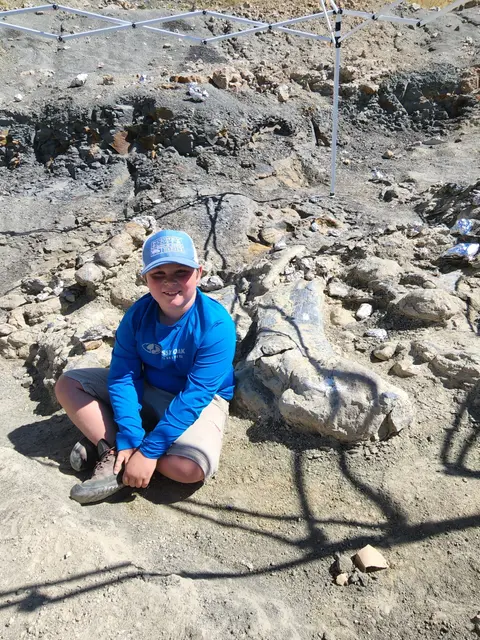 A young boy sitting next to a fossilized bone embedded in the ground.