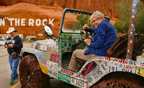 A smiling person sits in an artistic vehicle covered in license plates.