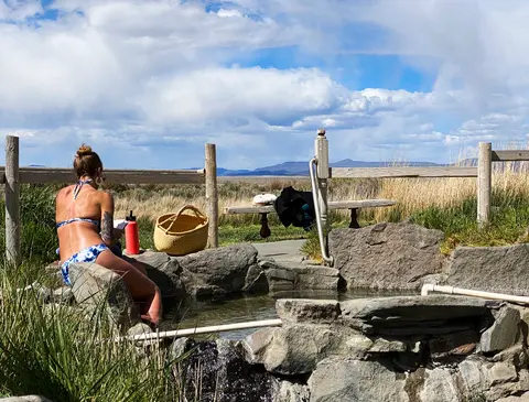 A woman sits by an outdoor rock pool overlooking a landscape under a cloudy sky.