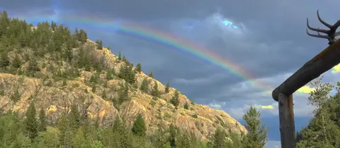 A vibrant rainbow arches over a steep, rocky, tree-covered mountainside under a cloudy sky.