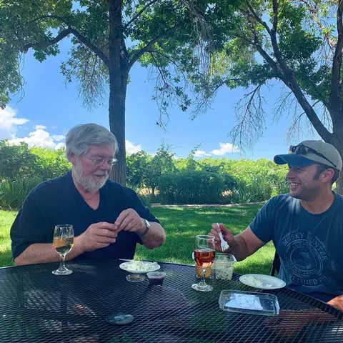 Two men sitting at a patio table outdoors with wine and food.