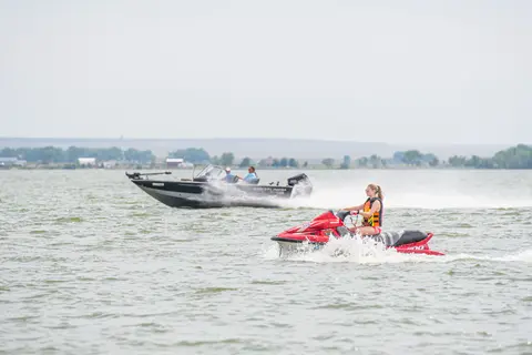 A person rides a personal watercraft on a lake with a boat in the background.