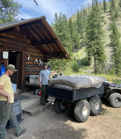 Two men stand outside a rustic log cabin next to a multi-wheeled trailer carrying mattresses.