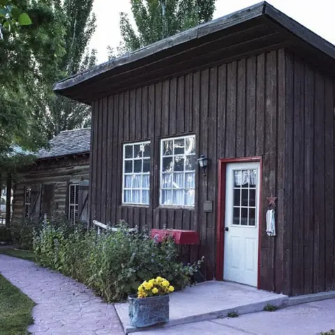 A rustic dark wood building with a white door and windows, alongside a stone walkway and garden.