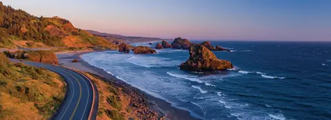 A winding coastal highway runs alongside a rocky ocean shore at sunset.