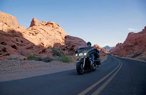 A person rides a motorcycle on a winding road through a desert landscape with red rock formations.