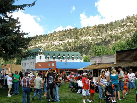 A crowd of people gathers on a grassy area in front of buildings and a rocky hillside.