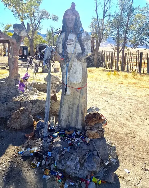 A statue stands outdoors in an arid landscape with trees and a wooden fence.