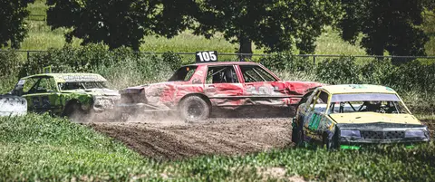Three damaged stock cars race on a dirt track.