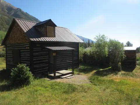 A dark wooden cabin with a metal roof sits in a grassy clearing with mountains in the background.
