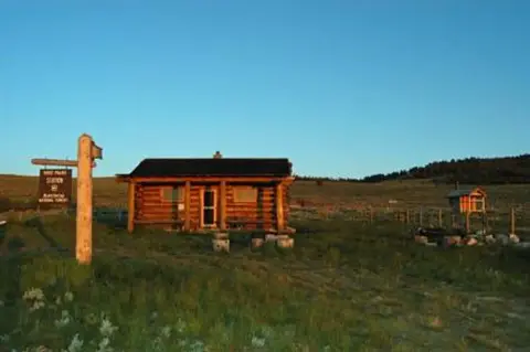 A log cabin sits in a grassy landscape beneath a blue sky.