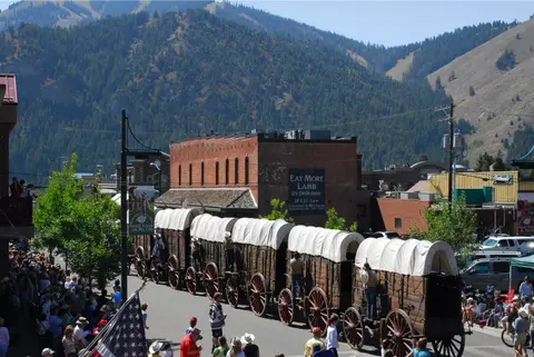 A line of covered wagons moves down a town street with mountains in the background.