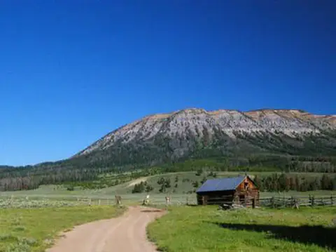 A dirt road leads past a small cabin toward a large mountain under a blue sky.