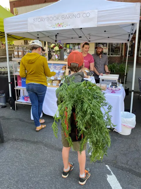 A child with a large bunch of leafy greens in a backpack stands in front of a market tent.