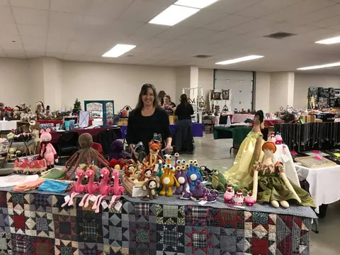 A woman stands behind a table covered in handmade dolls and toys at an indoor craft fair.