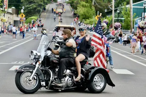 Two people ride a three-wheeled motorcycle in a parade on a street.