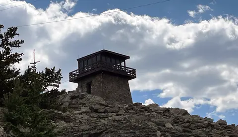 A stone and wood tower sits atop a rocky hill against a cloudy blue sky.