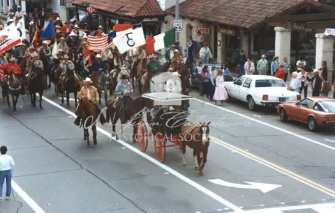 A parade featuring people on horseback and a horse-drawn carriage moving down a city street.
