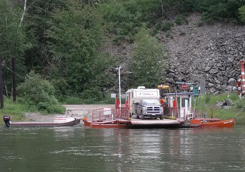 A small orange ferry carrying a truck and a camper on the water.