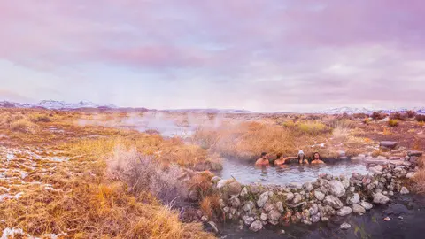 People soak in a natural hot spring surrounded by a landscape under a purple sky.