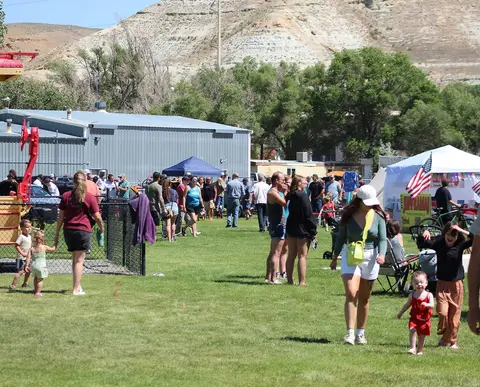 People walking and gathering on a grassy area outdoors near buildings and hills.