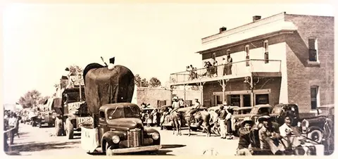 A historic photo showing a street scene with a parade float, cars, horses, and people gathered outside a building.