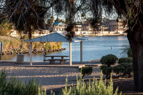 A shaded picnic shelter sits by a body of water with buildings in the distance.
