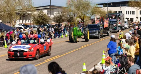 A parade featuring a decorated red car, a green tractor, and a pickup truck moves down a street lined with spectators.