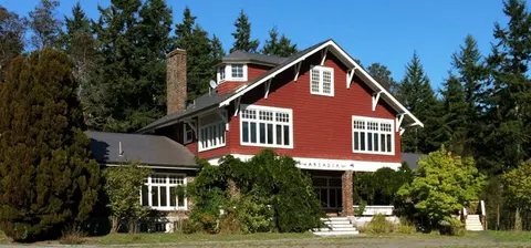 A red and white two-story house stands surrounded by tall green trees on a clear day.
