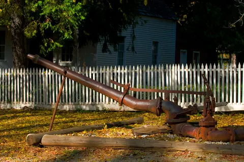 An old rusty pipe apparatus sits on the ground in front of a white fence and a building.