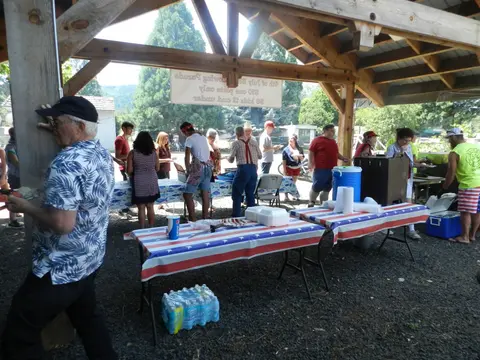 People gather under a wooden pavilion at an outdoor community event with food tables.
