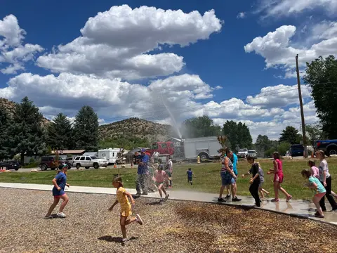 People and children playing in water sprayed from a fire truck outdoors on a sunny day.