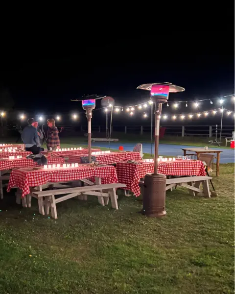 Picnic tables with red and white checkered tablecloths are set up outdoors at night with heaters and string lights.