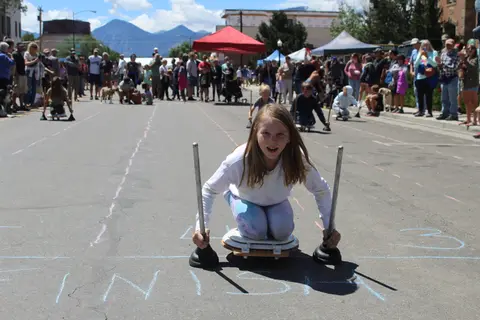 A young girl races a cart with plungers down a marked street during an outdoor event.