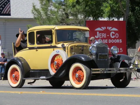 A vintage yellow and black car drives down a street with people waving.