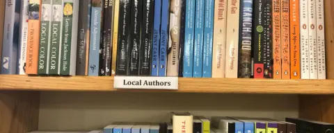A wooden bookshelf displaying various books with a local authors sign.