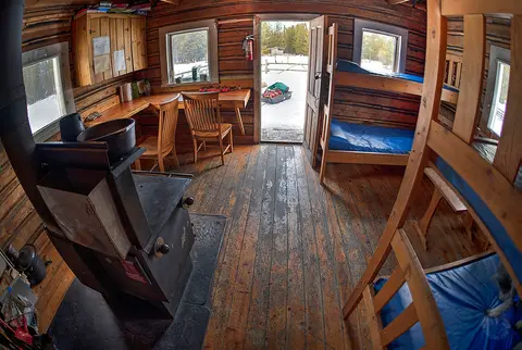 An interior view of a rustic log cabin with bunk beds, a wood-burning stove, and a desk.