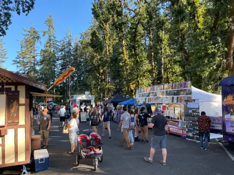 A crowd of people enjoying an outdoor market with food trucks and stands among tall trees.