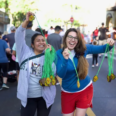 Two women smiling and holding up many medals on green lanyards outdoors.