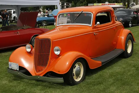 An orange classic hot rod parked on the grass at an outdoor car show.